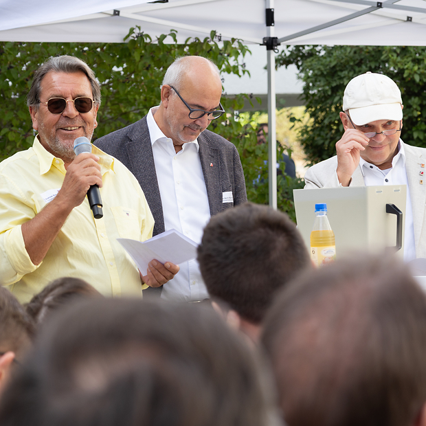 Drei ältere Herren unter einem Sonnenschirm vor einem Publikum.