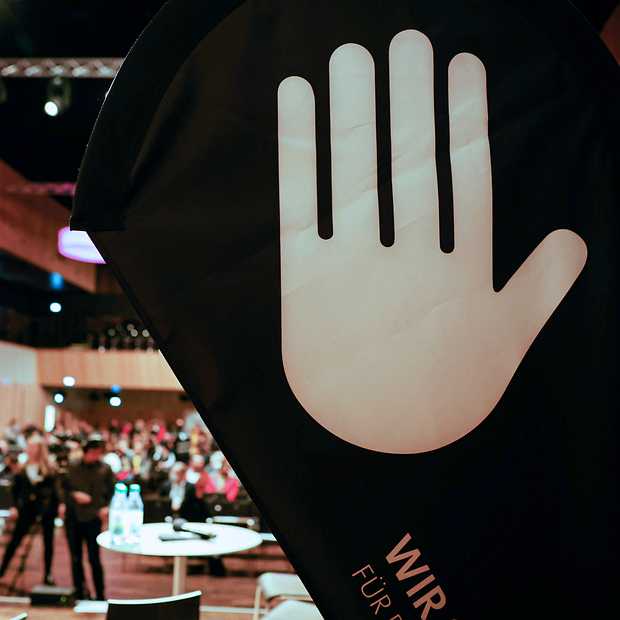 Grafik von erhobener Hand auf Rollup, im Hintergrund Halle innen mit Menschen