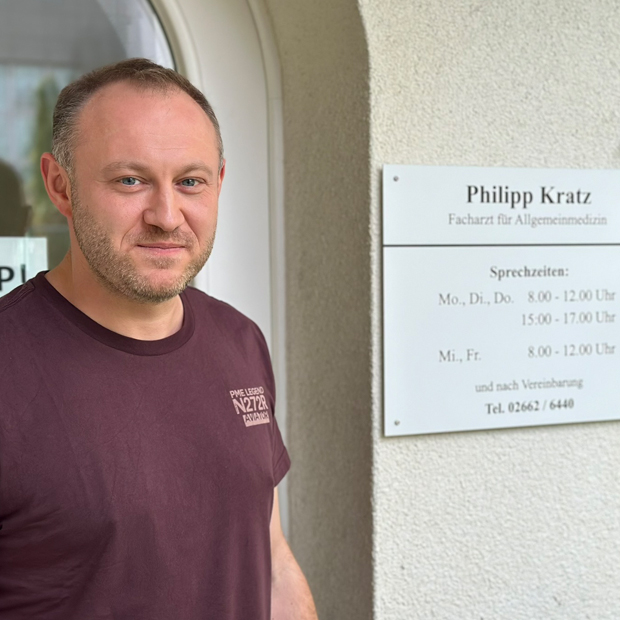 Philipp Kratz Mann mittleren Alters, lichtes Haar, im T-Shirt vor seiner Praxis, rechts daneben das Praxisschild