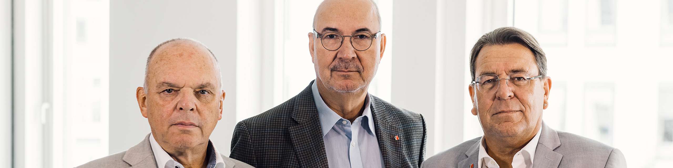 3 ältere Herren in Anzug, nebeneinander stehend, blicken ernst in die Kamera.