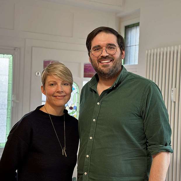 Dr. Melanie Galliardt und Christophe Mergeai Frau und Mann mittleren Alters, lächelnd, mit kurzen Haaren, leger gekleidet, er mit Bart und Brille