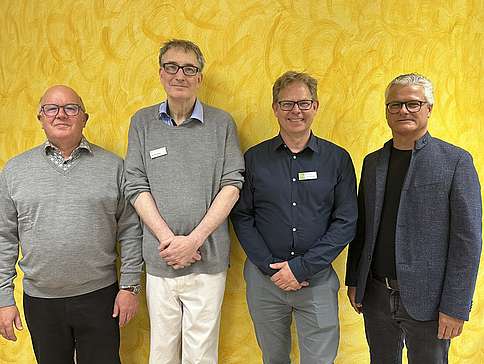 Vier ältere Männer, nebeneinanderstehend vor einer Wand, sie lächeln und blicken in die Kamera