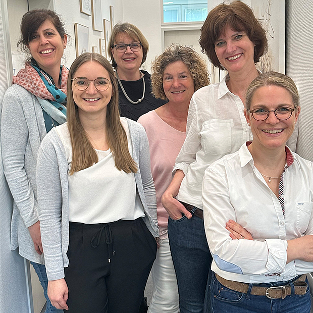 Dr. Manuela Käfer (r.) und Martina Hahn (2.v.r.) und Team 6 Frauen jungen und mittleren Alters in legerer Kleidung, stehend in Praxisflur, herzlich lächelnd