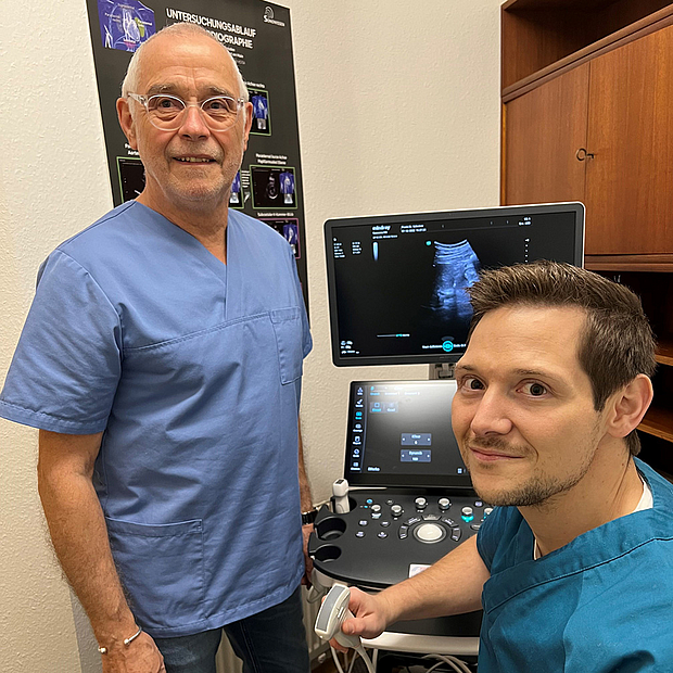 Dr. Jakob Schreiner mit Vater Dr. Jakob Schreiner sitzt vor einem Ultraschallgerät, links steht sein Vater, beide in OP-Kleidung