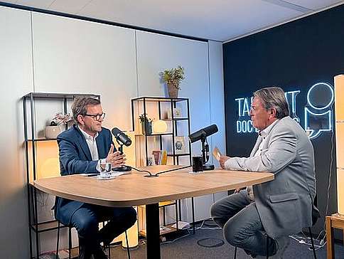 Ein Mann mittleren Alters und ein älterer Herr sitzen im Gespräch an einem Hochtisch.