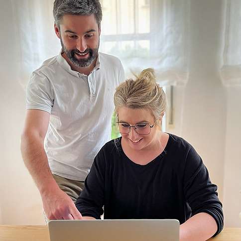 Dr. Philipp Rohleder mit Frau Franziska Frau und Mann mittleren Alters, sie mit Brille und Dutt sitzend am Laptop, er stehend. Sie lächeln.