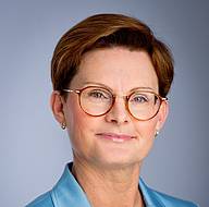 Zu sehen ist ein Portrait der Allgemeinmedizinerin Dr. Barbara Römer. Sie ist eine Frau mittleren Alters, hat rote-bräunliche kurze Haare und trägt eine Brille. Sie hat eine Bluse mit Blumen-Muster an und trägt ein hellblaues Sakko darüber. Dr. Römer lächelt freundlich in die Kamera.